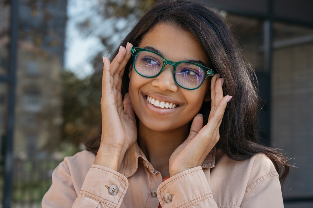 A smiling woman wearing green-rimmed glasses adjusts her frames while standing outdoors - Alpine Optometry | Optometrist Portola Valley A smiling woman wearing green-rimmed glasses adjusts her frames while standing outdoors - Alpine Optometry | Optometrist Portola Valley